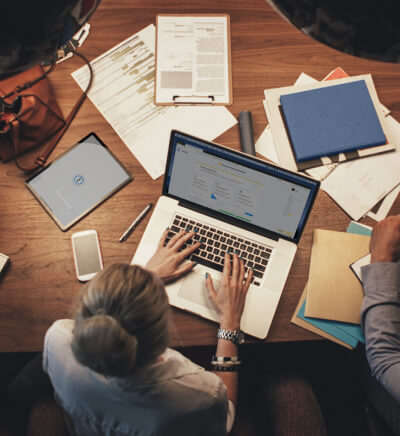 Aerial view of a blonde woman on her MacBook and a man sitting at a table surrounded by scattered papers and mobile devices